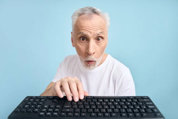 60多岁的老人惊奇地手牵着电脑电脑键盘 惊讶地表示着 转动着眼睛 张着嘴 在孤立的蓝色背景下摆出姿势 大家好技术 笔记本电脑Pov — 图库照片