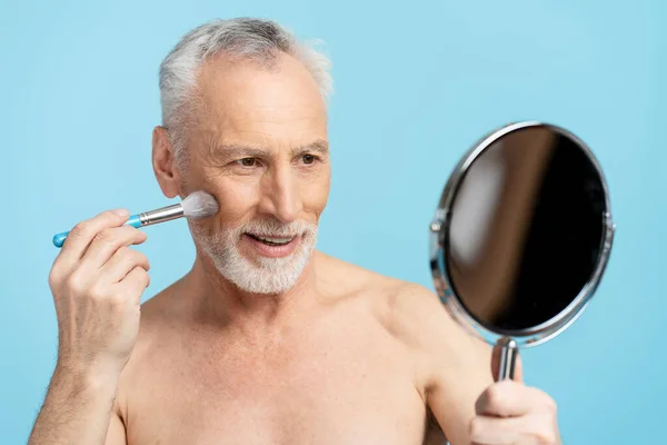 Smiling handsome elderly bearded man holding mirror doing makeup with makeup brush, looking in mirror isolated on blue background. Attractive man with naked torso after shower in bathroom