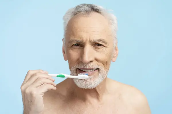 Attractive senior man looking in mirror, brushing teeth with toothbrush isolated on blue background. Concept of dental care and health