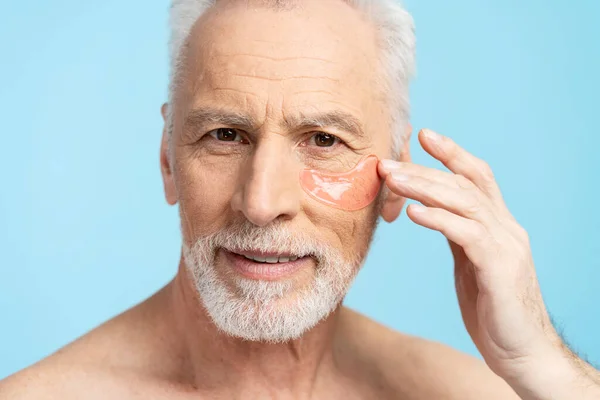Closeup portrait of gray haired man applying patches, skin care isolated on blue background. Elderly male looking in mirror, morning routine