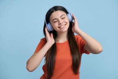 Beyaz, mutlu, parlak turuncu tişörtlü genç kız, geniş gülümsemeli, kulaklıkla güzel bir müzik dinleyerek eğleniyor, mavi arka planda izole edilmiş. - İnsanlar. Yaşam tarzı. Eğlence