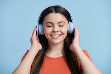 Yakın plan güzel genç kız mavi kablosuz kulaklıklardan müzik dinliyor, havalı film müziğinin keyfini çıkarıyor, dişlek bir gülümsemeyle gülümsüyor, gözleri kapalı mavi renkli stüdyo arka planında poz veriyor.