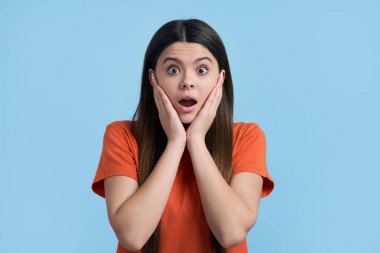 Caucasian lovely teenager girl putting hands on her face, expressing amazement and frustration, looking at camera, isolated over blue color background. Adolescent child looking surprised, astonished clipart