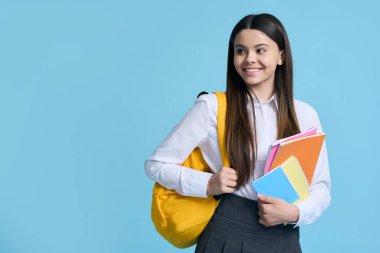 Mutlu beyaz kız, sarı sırt çantalı ve ders kitaplı liseli kız, yan bakıyor, mavi arka planda soyutlanmış. - İnsanlar. Eğitim. Akademik yılın yeni döneminde okula dönüyorum.