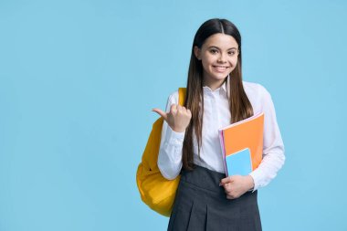 Sıradan okul üniforması giymiş, sarı sırt çantası ve kitaplarla poz veren, kameraya gülümseyen, mavi izole edilmiş arka planda reklamlar için fotokopi alanı gösteren çekici, zeki ve genç bir kız öğrenci.