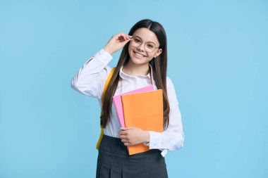 Beyaz, güzel, pozitif, zeki, gözlüklü, günlük okul üniforması giyen, ders kitaplarını tutan, şık gözlükleriyle kameraya gülümseyen, izole edilmiş mavi arka plan.