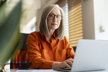 Kendine güvenen olgun bir iş kadınının portresi şık gözlükler takan, turuncu gömlekli modern ofiste çalışan dizüstü bilgisayarı kullanan. Başarılı yönetici planlama projesi, klavyede yazma