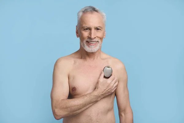 Happy senior man holding a pacemaker over his naked torso, smiling, confidently looking at camera, standing shirtless, isolated on blue background. Healthcare and medicine. Cardiac surgery concept
