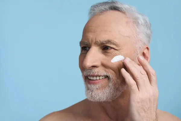 Headshot of a Caucasian handsome senior man, applying moisturizing nourishing rejuvenating cream on his face, isolated over blue color background with copy space. Skin care. Male beauty. Cosmetology