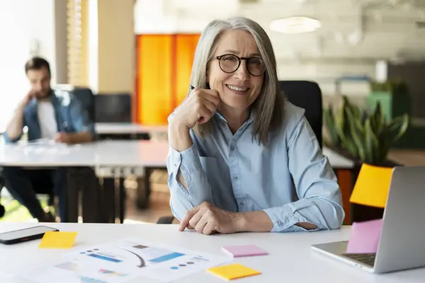 Portrait of smiling confident senior businesswoman using sticky notes looking at camera sitting in modern office. Gray haired manager wearing stylish eyeglasses at workplace. Scrum, agile concept