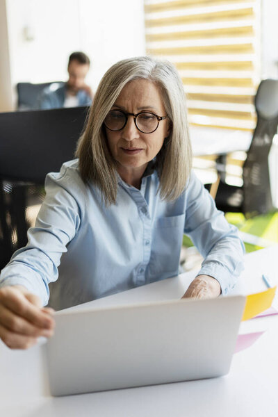 Кавказка уверенная в себе элегантная седовласая зрелая деловая женщина, в повседневной одежде и очках, сидящая за столом с ноутбуком, работающая в современном офисном интерьере. Командная работа. Корпоративный бизнес. Рабочий день