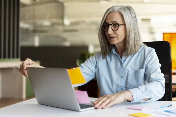 魅力的なグレーハイレッドシニアビジネス女性は ラップトップコンピュータを使用して眼鏡を着用し 現代のオフィスでオンラインで作業し 粘着性のあるノートを使用して成功したマネージャー アジャイル方法論 スクラムコンセプト — ストック写真