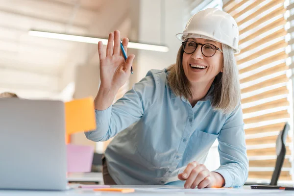 Smiling mature woman in helmet waving with her hand, standing at desk, planning new design project, in modern office interior. Portrait of senior female architect, design engineer at workplace