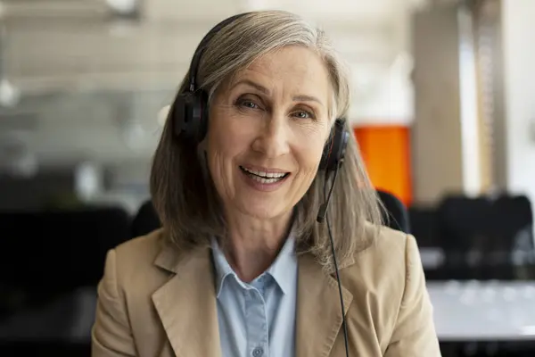 Portrait of smiling senior woman, call center operator answering call, communication with customer sitting in modern office. Serious freelancer having video call working from home. Successful business