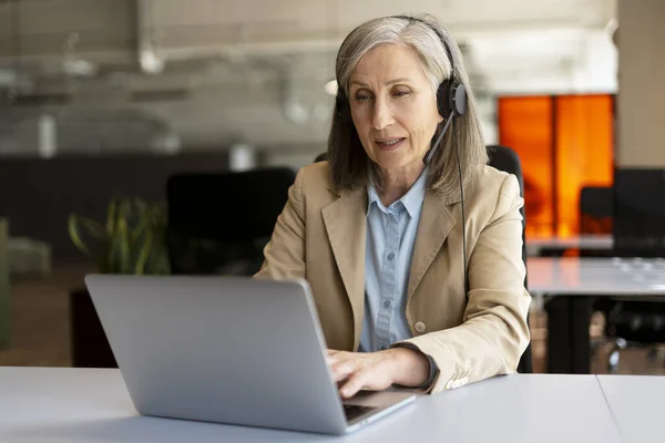 Senior woman, call center operator using laptop answering call, communication with customer sitting in modern office. Serious freelancer having video call working from home. Successful business