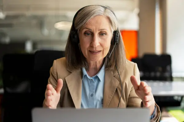 Senior woman, call center operator using laptop, answering call, communication with customer sitting in modern office. Serious freelancer having video call working from home. Successful business