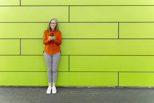 Portrait of smiling grey haired senior woman holding mobile phone communication online, reading text message standing near green wall, copy space. Stylish female using smartphone watching video