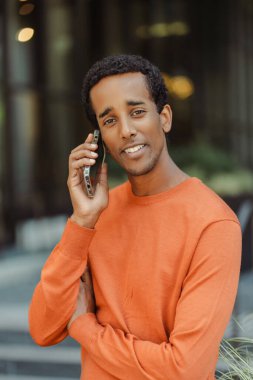 Yakışıklı, gülümseyen Afro-Amerikan bir adamın portresi. Cep telefonuyla konuşan, telefona cevap veren, şehir sokağına bakan. Başarılı iş, teknoloji kavramı 