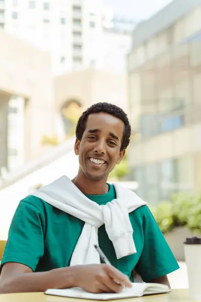 Smiling african american man, writer sitting at desk in cafe, taking notes in diary looking at camera outdoors. Handsome student preparing for exam on university campus