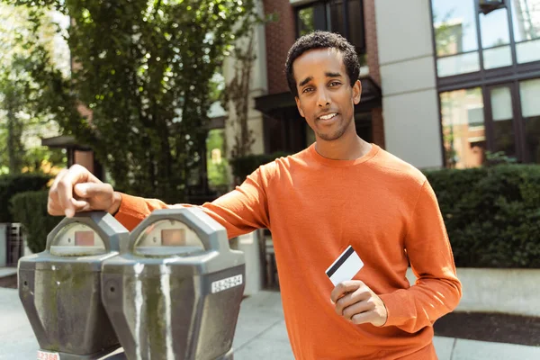 Portrait of attractive smiling African American man standing at parking meter, holding credit card, paying for parking, looking at camera outdoors. Transportation concept