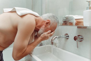 Havlulu, beyaz saçlı emekli bir adamın portresi, çamaşır leğeninde çıplak bir gövde, musluktan akan su, apartmanın banyosu. Hijyenik prosedür kavramı, sabah rutini