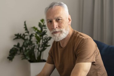 Kahverengi t-shirt giyen, oturma odasındaki koltukta oturan, arka planda saksı saksısı olan yakışıklı emeklinin portresi. Çekici son sınıf öğrencisi fotoğraf için poz veriyor, kameraya bakıyor.