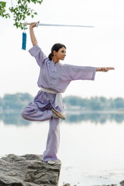 Kung fu ustası kadın, elinde kılıç tutan kimono giyiyor, suyun yanında taşın üzerinde Wushu çalışıyor, kopyalama uzayı. Sağlıklı yaşam tarzı, dövüş sanatları konsepti