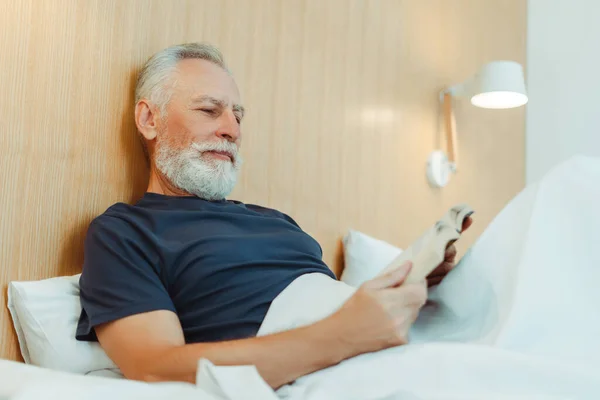 一个面带微笑的白发苍苍的老人的画像 躺在床上 上面铺着毛毯 手里拿着一本书 在时尚的公寓里看书 英俊而成熟的男性在酒店休息 — 图库照片