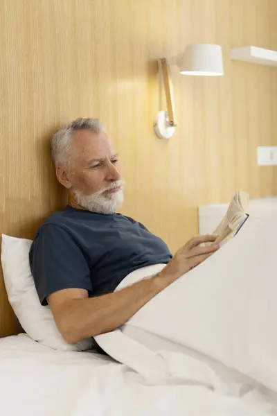 Portrait of smart elderly man lying in bed covered with blanket holding book, reading in stylish apartment. Handsome mature male resting in hotel