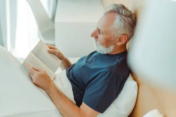 Attractive elderly man lies in bed on pillow, covered with blanket, reading book, relaxing in living room. Smart bearded grandfather resting. Retirement concept