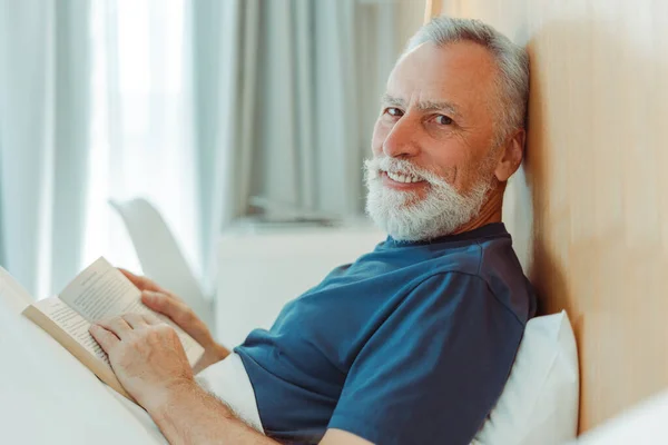 Portrait of smiling gray haired elderly man lying in bed covered with blanket holding book, reading in stylish living room, looking at camera. Handsome mature male resting in hotel