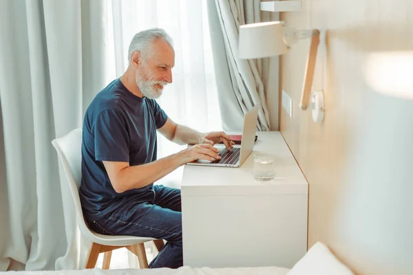 积极微笑的老年人坐在客厅 宾馆的办公桌前 用笔记本电脑 年长的复印机打字 英俊的祖父玩网络游戏 技术概念 — 图库照片