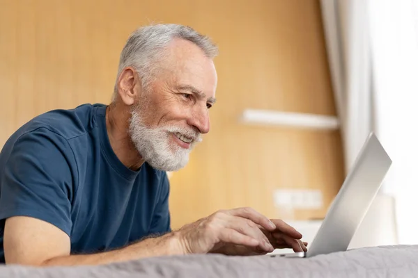 带着胡子自信微笑的成熟男人的画像 躺在酒店的床上 成功的老年养恤金领取者在客厅玩游戏 在家工作的概念 — 图库照片