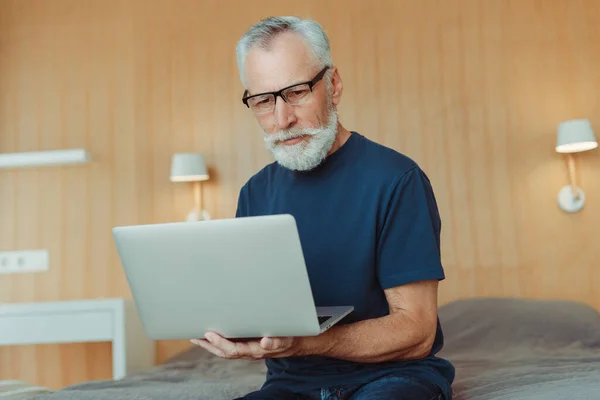 一个灰色头发的老人头戴眼镜 坐在客厅的床上 用笔记本电脑 打字机打字 远程工作的画像 专业的高级程序员在家里工作 技术概念 — 图库照片