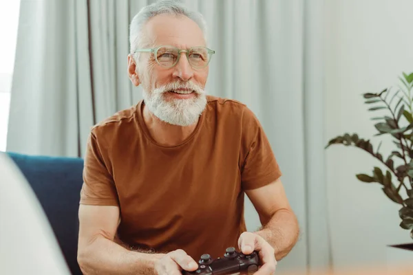 在公寓里坐在舒适的沙发上玩电子游戏 脸上挂着眼镜笑着 有胡子的积极祖父玩得很开心 技术概念 — 图库照片