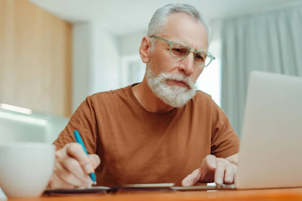 戴眼镜 坐在办公桌前 使用笔记本电脑 记笔记 在家工作的聪明而严肃的老年人的画像 有吸引力的退休人员在网上工作 远程工作概念 — 图库照片