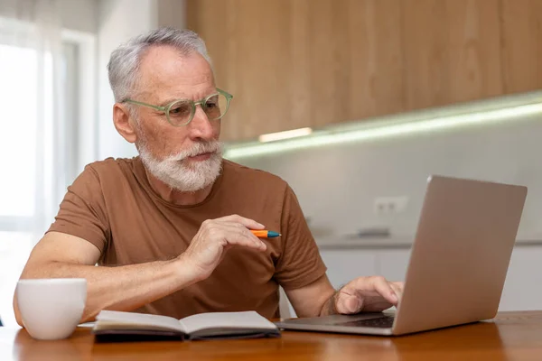 形象迷人 严肃的老年人戴眼镜 坐在办公桌前 使用笔记本电脑 记笔记 网上教育 在家工作 漂亮的退休人员在网上工作 远程工作概念 — 图库照片