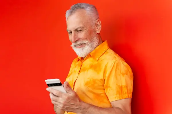 笑容可亲的老男人拿着手机看短信 网上购物被隔离在红色背景之下 留着胡子的嬉皮士在街上用智能手机和通讯移动银行概念 — 图库照片