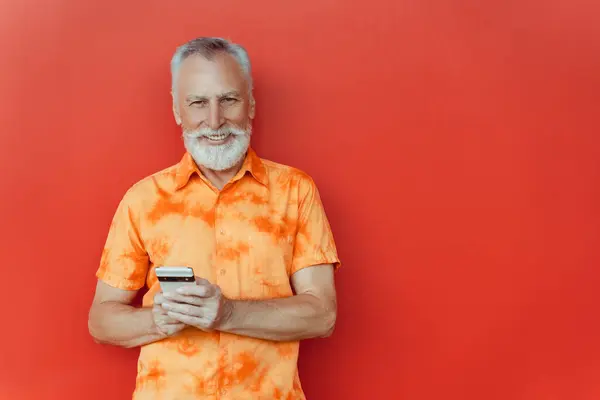 微笑的老人站在红墙边的街上 拿着手机在线购物 复制空间 快乐英俊的大胡子嬉皮士使用智能手机 在线交流 移动银行 — 图库照片