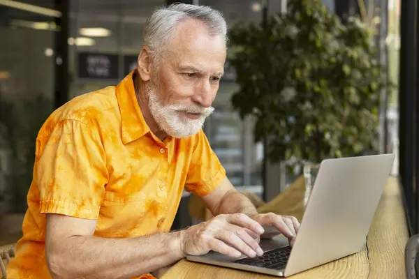 一个有魅力的老年人的画像 一个使用笔记本电脑的自由职业者 坐在咖啡店的工作场所 网上穿着休闲装的英俊的退休人员 打字的抄写员 技术概念 — 图库照片