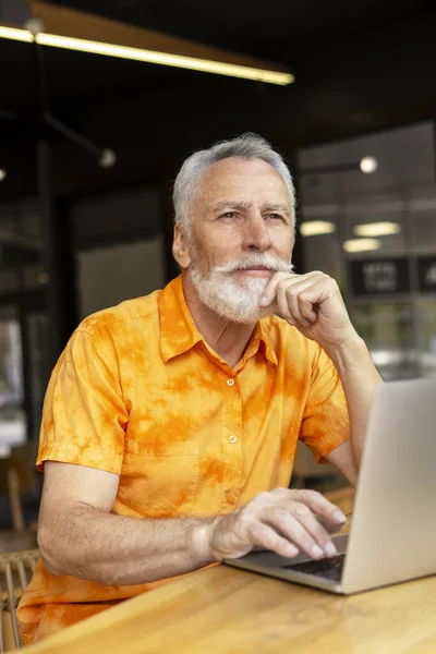 一个多愁善感的老人 用笔记本电脑写作 坐在工作场所 上网工作 迷人的胡子养恤金领取者 时髦的嬉皮士网上购物 看着别处 技术概念 — 图库照片