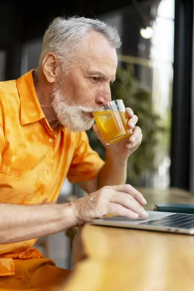 身着夏装的退休男子身着休闲装 手持笔记本电脑 观看视频 喝鸡尾酒 坐在咖啡店里 迷人的老年男性远程工作 网上购物 成功的假期概念 — 图库照片