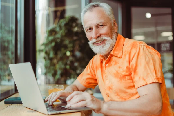 用笔记本电脑 在现代化的办公室里工作 坐在办公室里 穿着橙色衬衫 满脸胡须的成熟男人 吸引退休自由职业者的远程工作 看着相机 业务概念 — 图库照片
