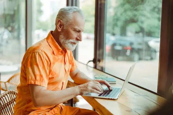 有魅力的老年人 穿着时髦休闲装的自由职业者 使用笔记本电脑 上网在电脑上工作 坐在靠窗的工作场所 老文案作者打字 远程工作的概念 — 图库照片
