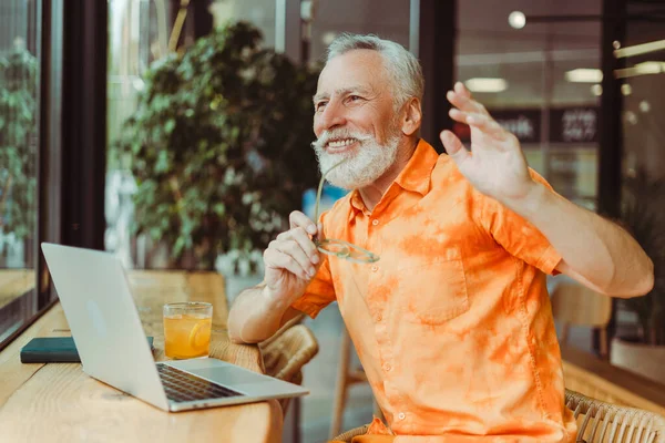 微笑英俊的老年人穿着时髦的橙色衬衫 举着眼镜 使用笔记本电脑 坐在工作场所 迷人的留胡子养恤金领取者坐在咖啡馆里度假 夏季概念 — 图库照片