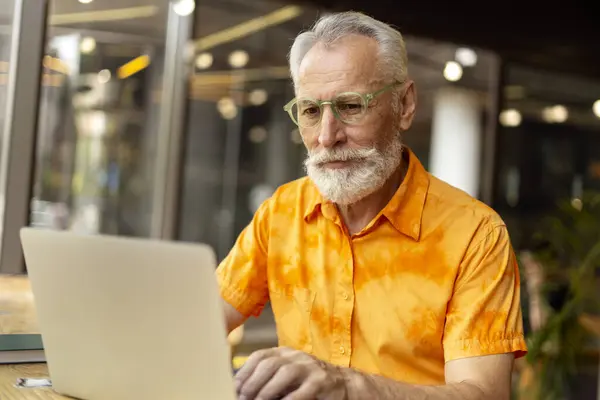 面带微笑的成熟自由职业者的画像 戴着时髦橙色衬衫的眼镜 使用笔记本电脑 坐在现代办公室或咖啡馆里 英俊的老年作家在计算机上打字 — 图库照片