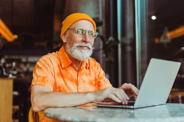 英俊的老人微笑着 留着胡子的嬉皮士戴着黄色的帽子 用笔记本电脑 坐在现代咖啡馆的网上工作 时尚商人计划创业 成功的商业概念 — 图库照片