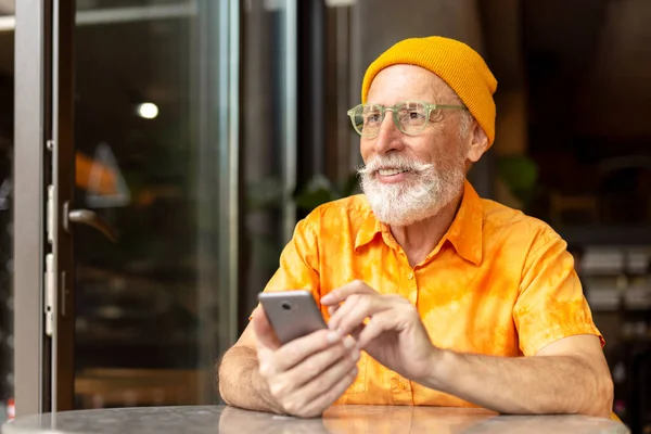 Smiling senior man, stylish bearded hipster holding mobile phone using modern app shopping online sitting in cafe. Happy handsome freelancer receive payment. check email. Technology, remote job
