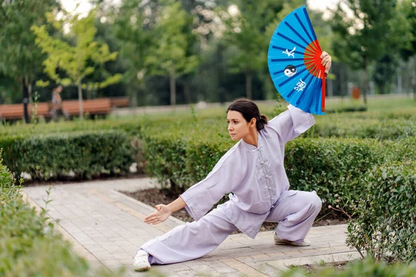 Caucasian woman with fan wearing kimono practicing wushu at park. Chinese martial arts, healthy lifestyle concept