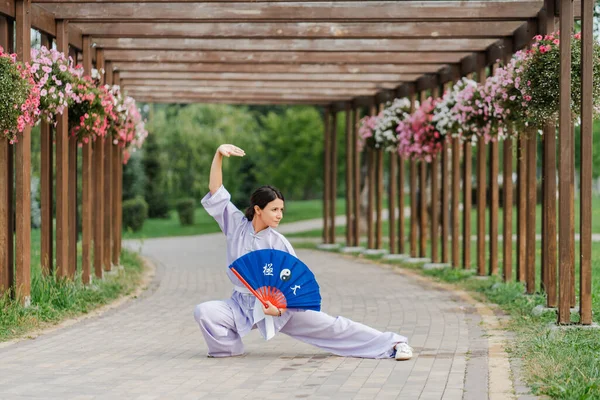 Caucasian woman with fan wearing kimono posing at at green park after practicing wushu. Chinese martial arts, healthy lifestyle concept